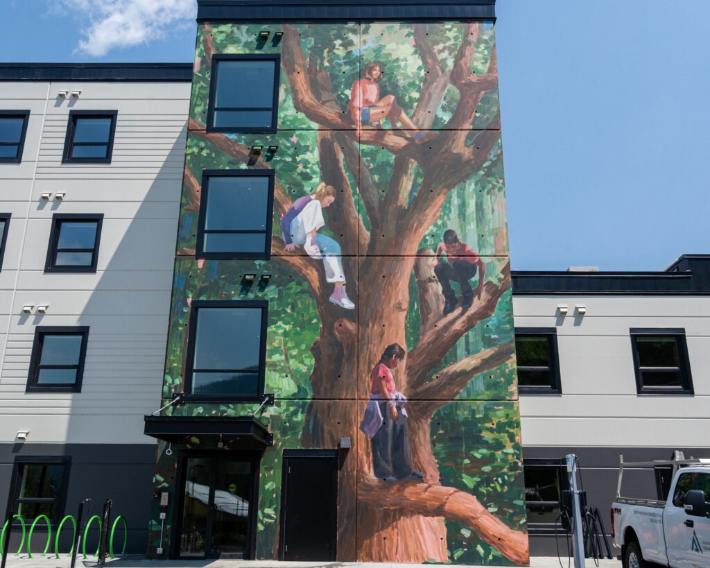 Among the trees by Megan Oldhues. A large tree with several children sitting in its branches. Green foliage covers the background of the mural.