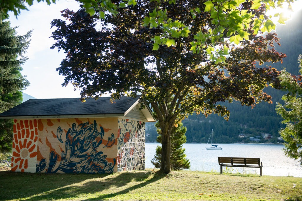 A small outbuilding covered in a wrap mural next to the lake, a large tree, and a park bench.
