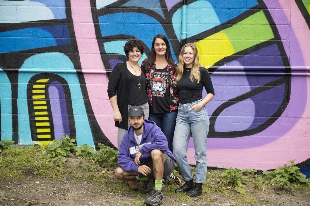Sydney Black, Amber Santos, Nicola Rough, and Andrew Tavukciyan stand in front of an abstract mural