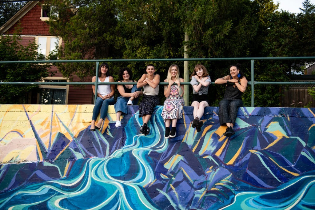 Six NIMF team members sit on the ledge of a colorful mural of mountains and rivers. From left to right: Amber Santos, Sydney Black, Lucy Brennan, Zan Comerford, Ingrid Love, and Dee Landsberg
